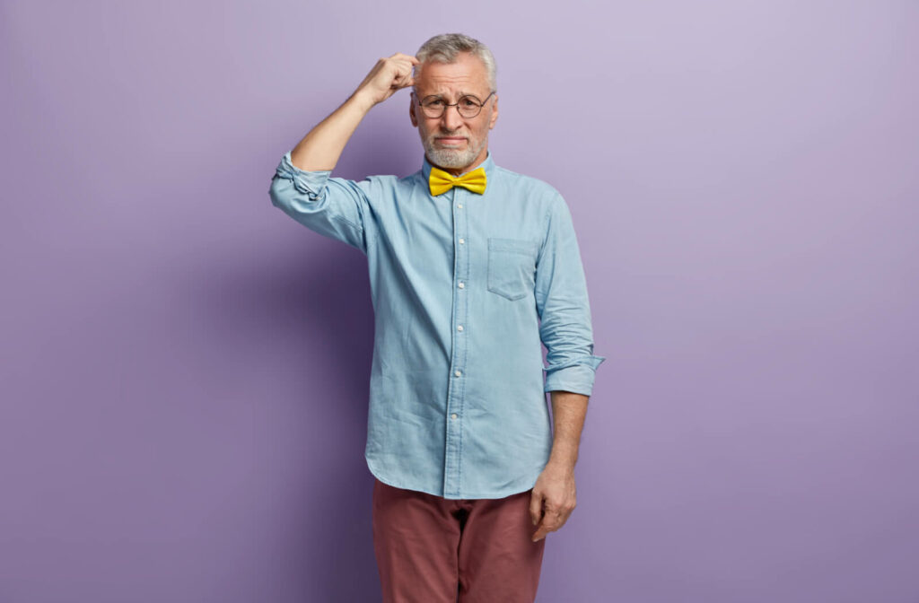 A senior man wearing a yellow bowtie and glasses scratching his head, confused, as he stands against a purple backdrop.