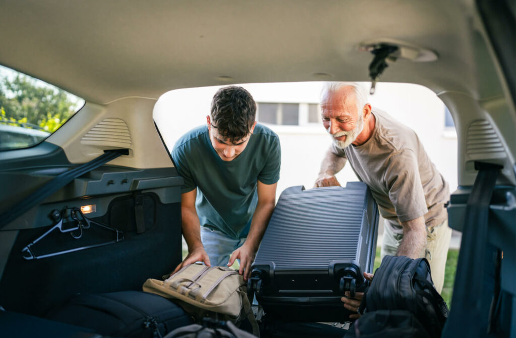 A young man helping his senior father put luggage inside a car.